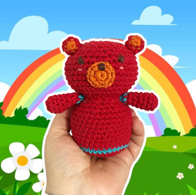 A crochet pattern for a red bear doll named Boba with a rainbow and flowers in the background.