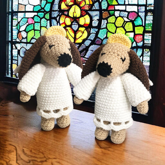 Two crocheted angel dog toys with beige, white, and brown colors, standing in front of a stained glass window.