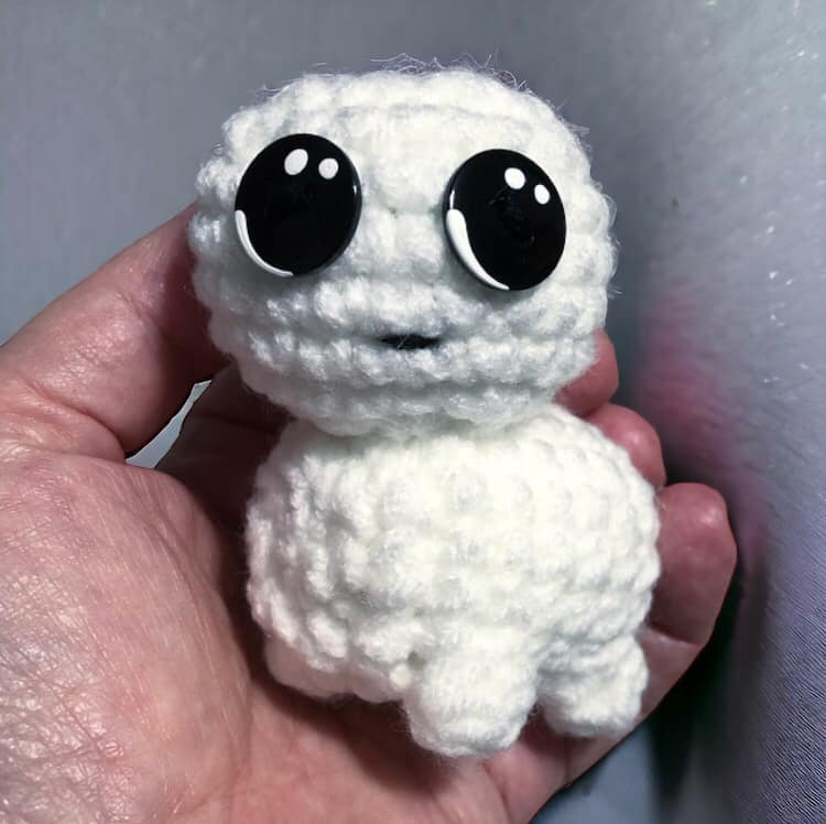 A crocheted version of the tbh creature being held up against a white wall.
