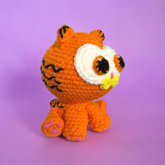 A crochet plush toy resembling the character Garfield, made from orange yarn with additional features in other colours against a purple background.
