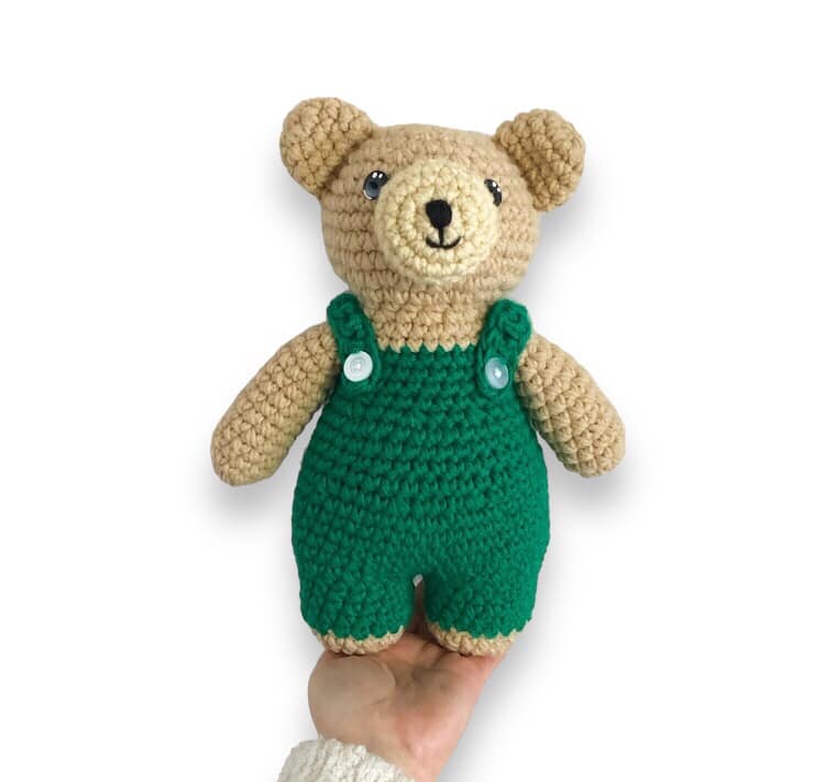 A crochet doll resembling Corduroy Bear being held up against a white background.