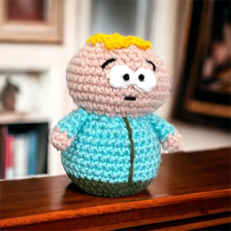A crochet version of Butters from South Park standing on a shelf with photos in the background.