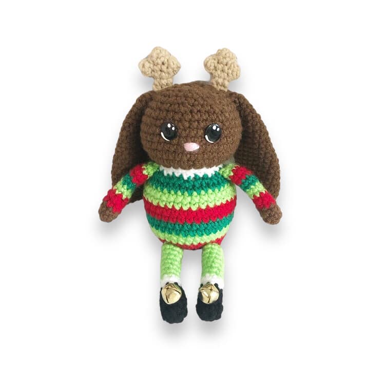 A crocheted jackalope doll with small antlers and a green and red striped sweater with little black boots that have bells on them sitting against a white backround.