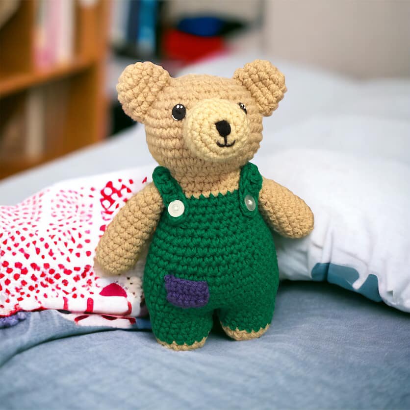 A crocheted doll resembling Corduroy Bear standing on a blue bed.