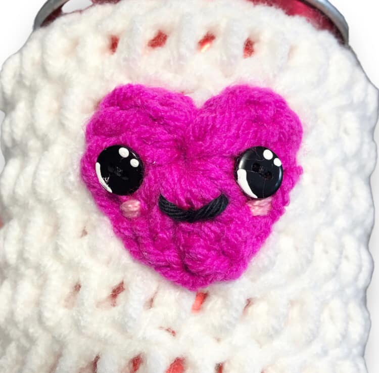 A close up of the cup cozy. It has a pink heart with large button eyes and a smiley face.