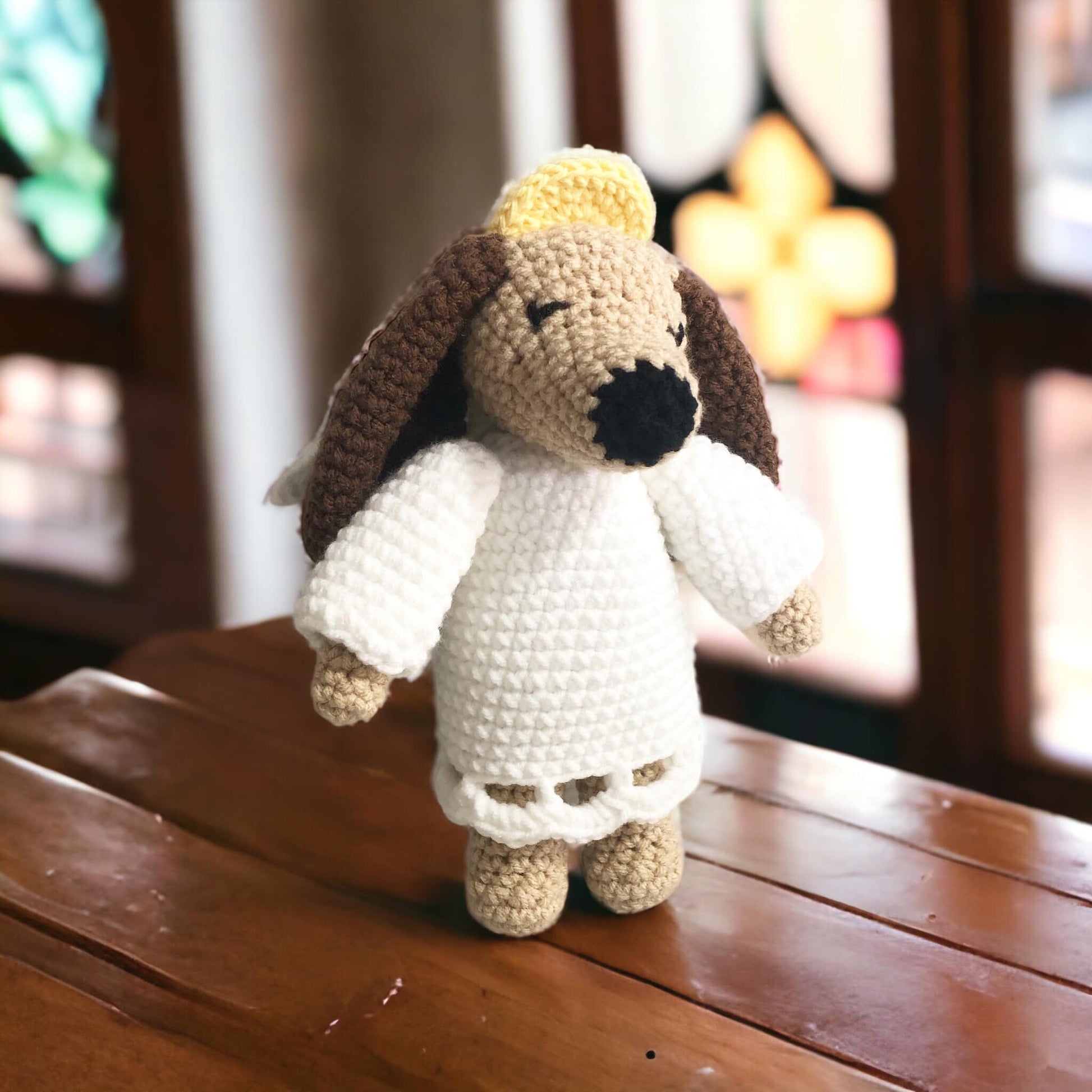 A crocheted angel dog toy with beige, white, and brown colors, standing in front of a stained glass window.