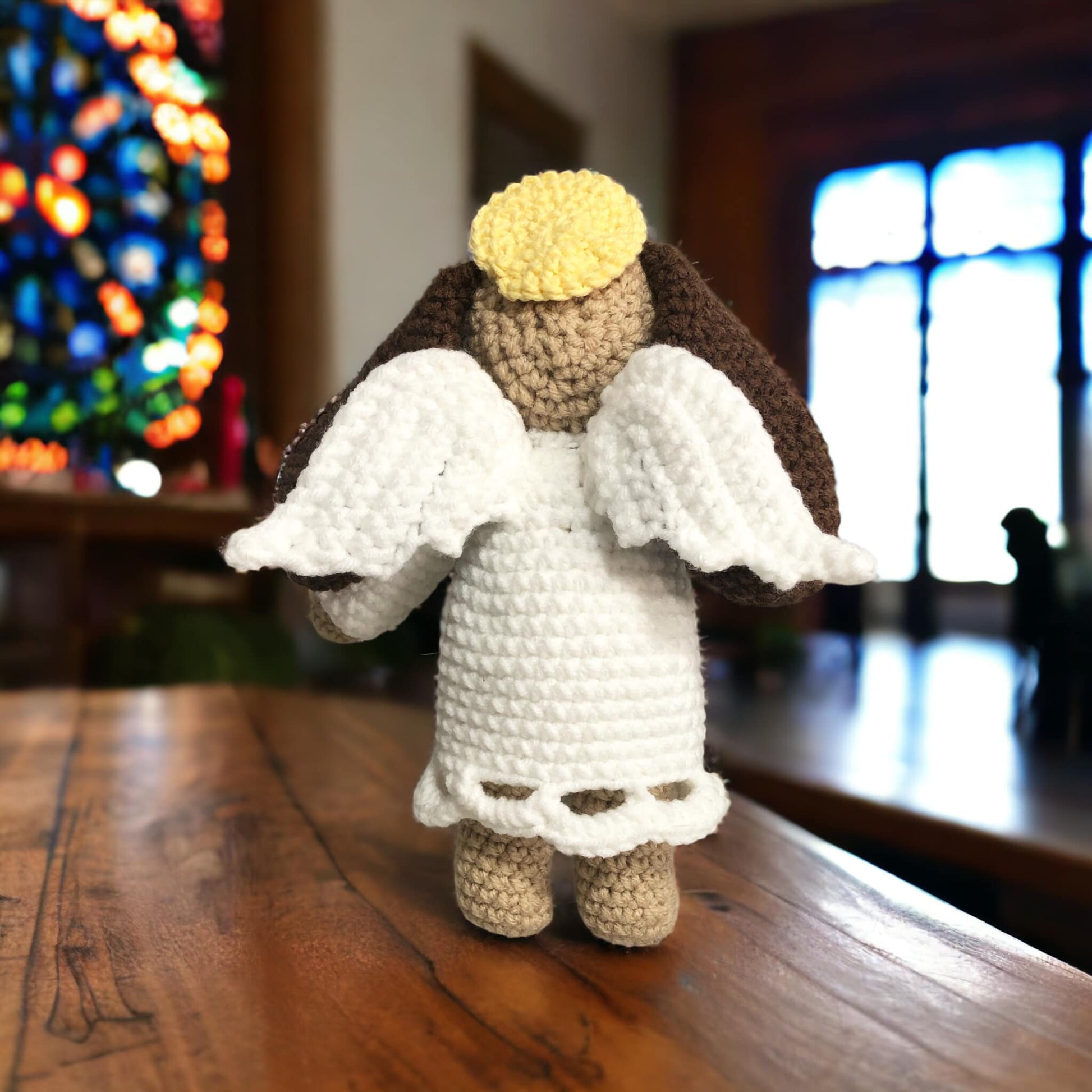 A crocheted angel dog toy with beige, white, and brown colors, standing in front of a stained glass window and cacing away from the camera showing off the wings.