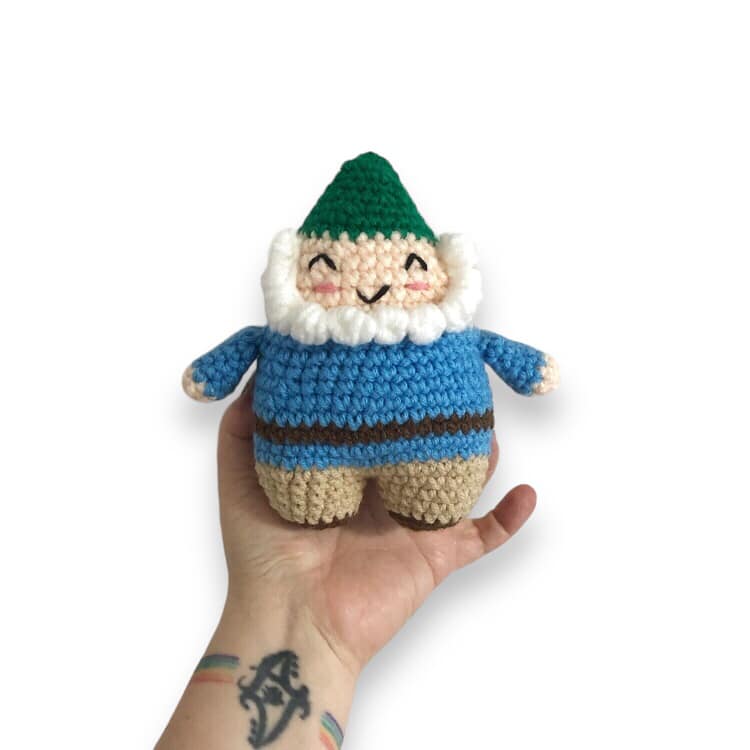 A crocheted doll of a smiling garden gnome with a green hat, blue jacket, beige pants, and a white beard being held up against a white background.