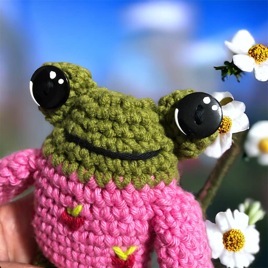 A crocheted frog doll in a pink sweater with strawberries against a background with white flowers.