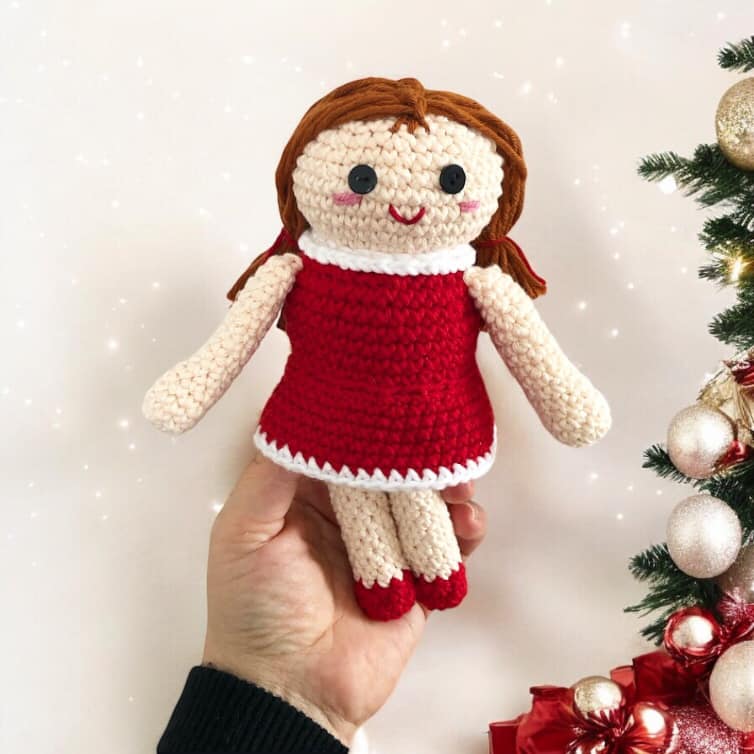 A crocheted version of the doll from misfit island being held up next to a Christmas tree.