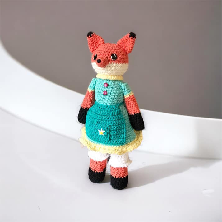 A crochet fox doll with a blue dress and bloomers standing against a white and lavender background.