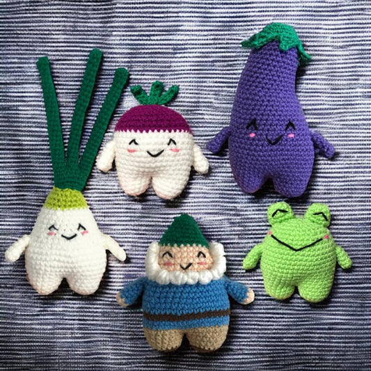 A series of crochet dolls each resembling an eggplant, a frog, a turnip, a green onion, and a gnome with a blue shirt and dark green hat against a blue and white stripe background.