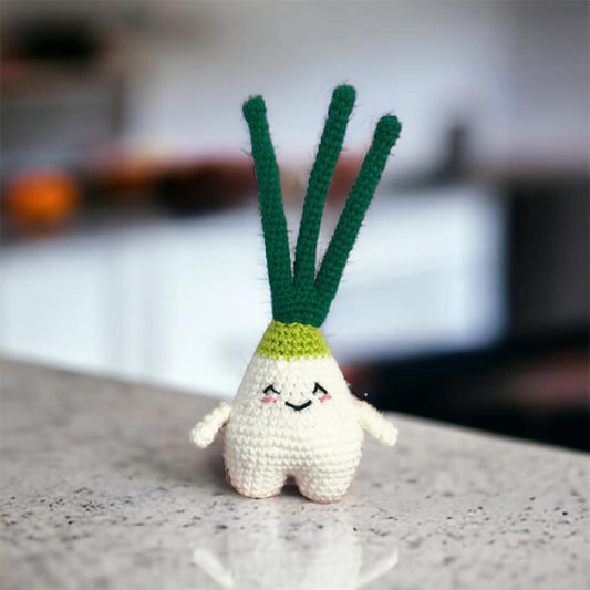 A crochet amigurumi toy resembling a green onion with a smiling face, featuring white body, green leaves, and black details standing on a white marble countertop.