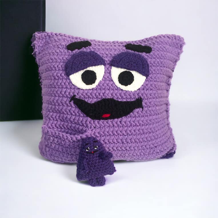 A crocheted Grimace pillow with Grimace pocket pal sitting against a white background.