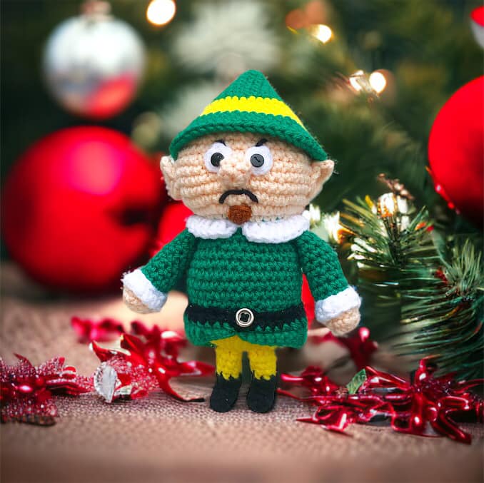 A crocheted grouchy elf from Rudolph the Red-Nosed Reindeer standing against a background of Christmas decor.