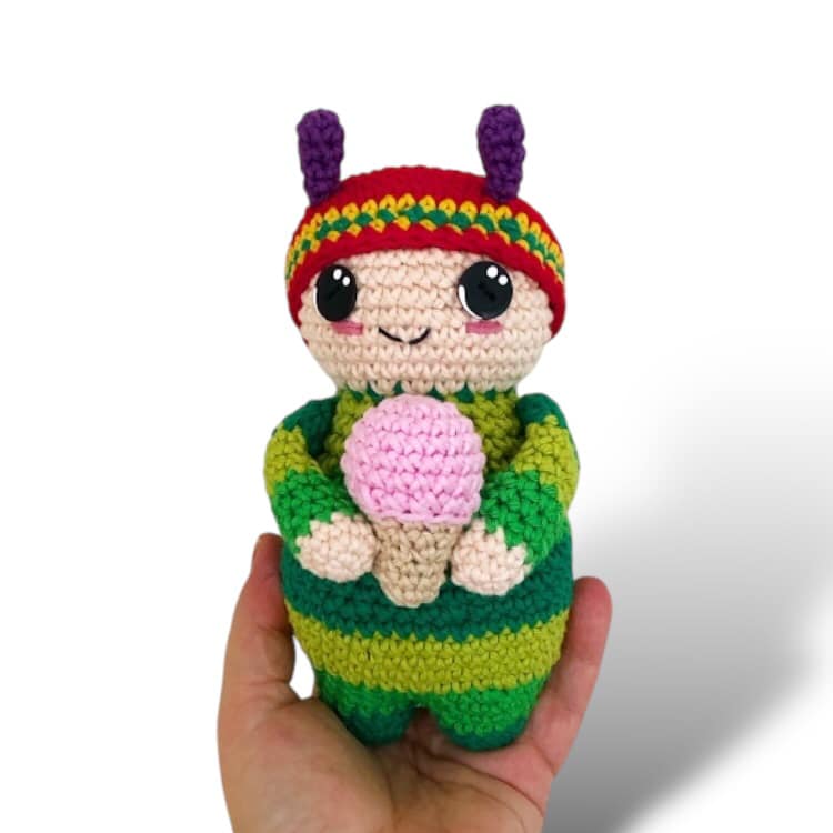 A crochet pattern for a toy resembling a small child dressed like caterpillar with a striped body, wearing a hat, and holding a pink ice cream cone. The image shows the finished toy.