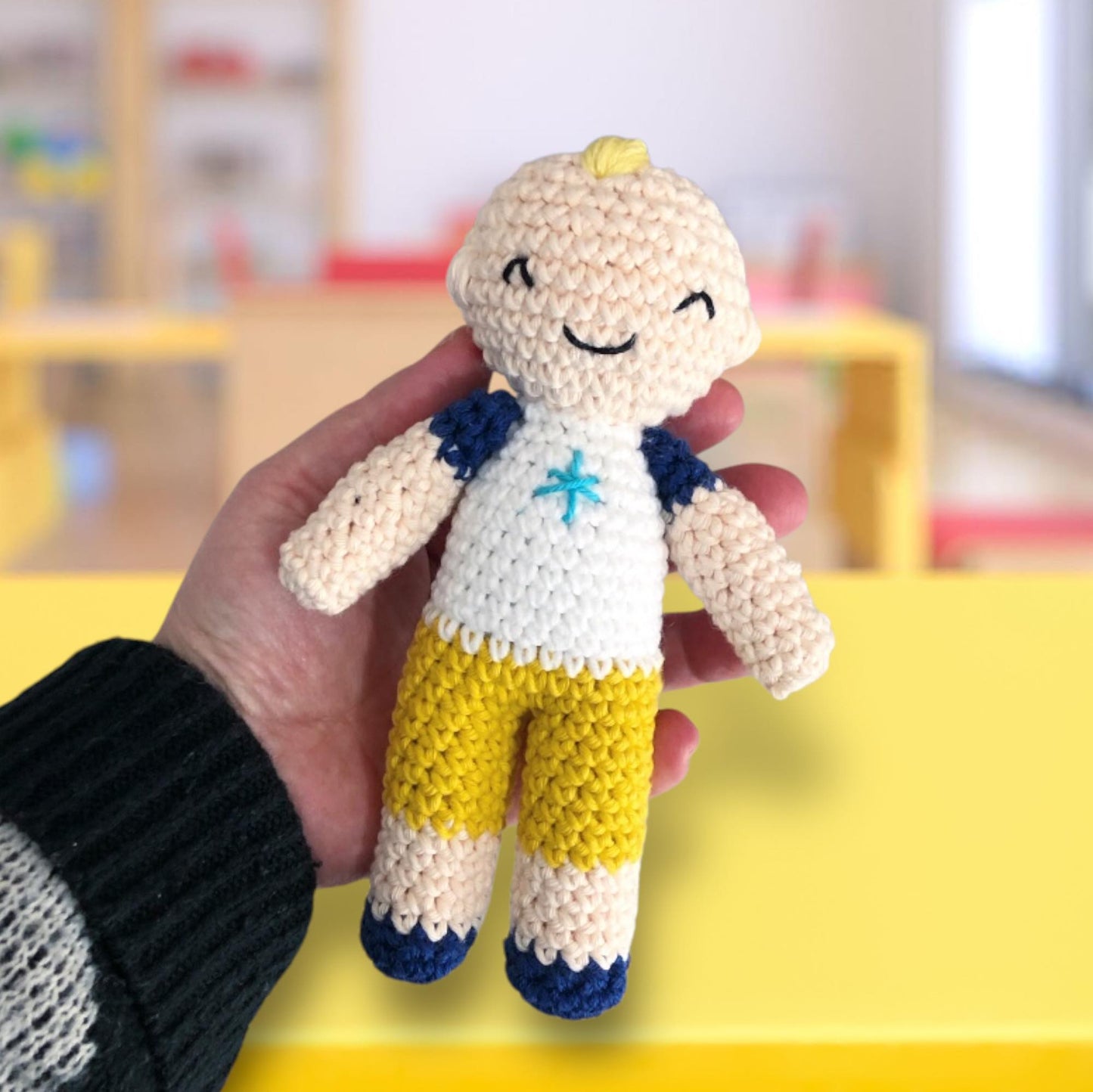 A crochet doll of JJ being held up against a backdrop of a school setting.