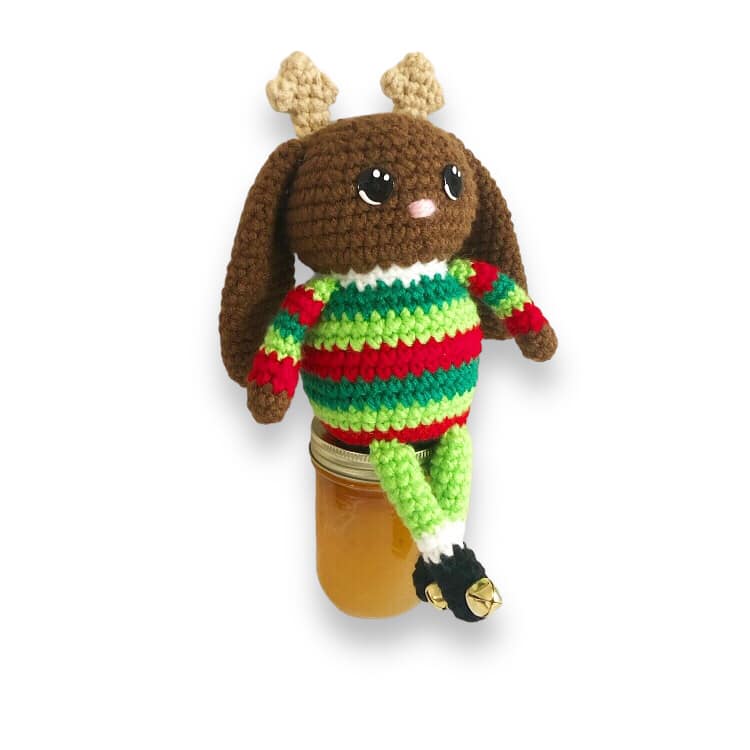 A crocheted jackalope doll with small antlers and a green and red striped sweater with little black boots that have bells on them sitting on a jar.