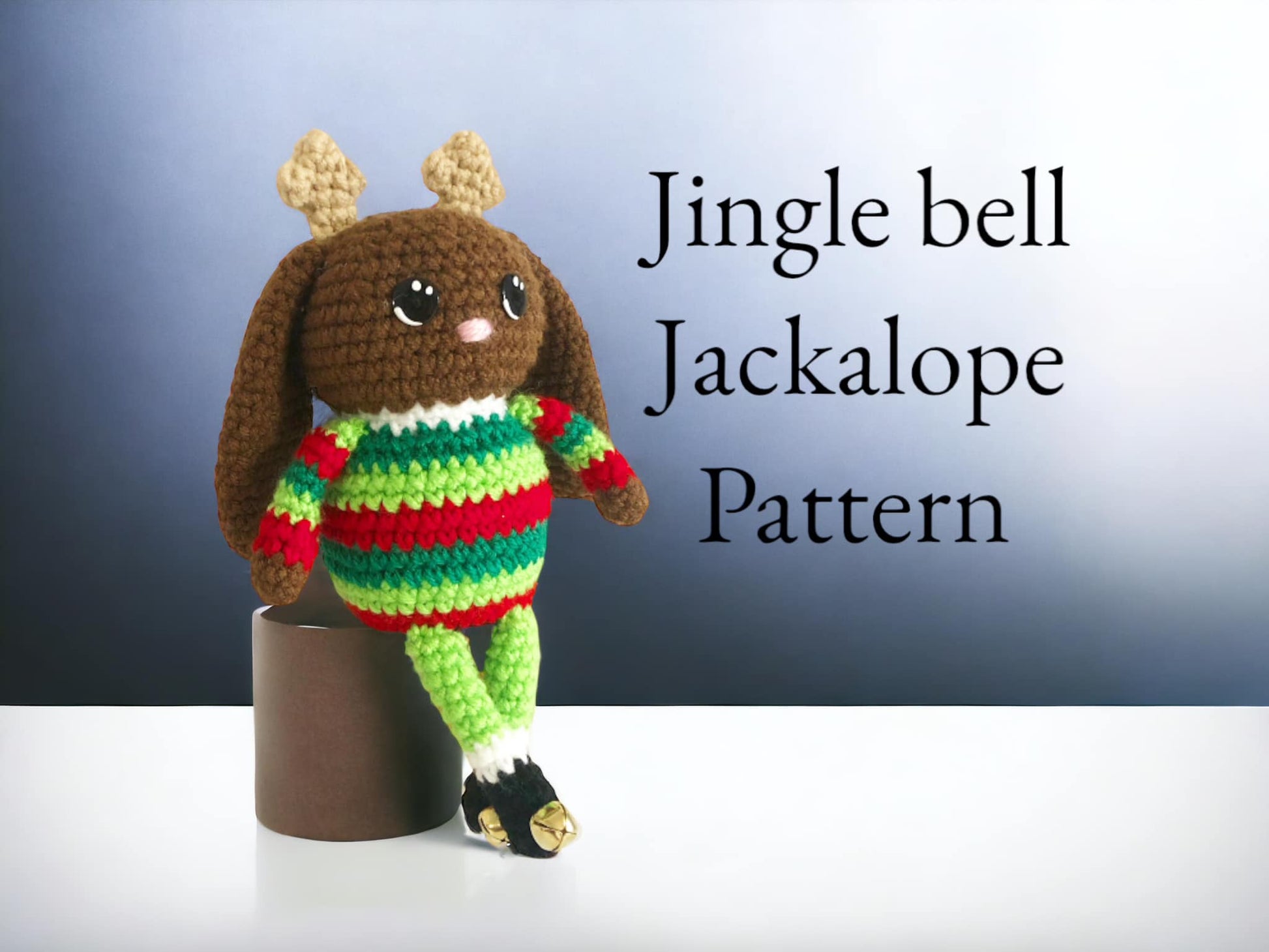 A crocheted jackalope doll with small antlers and a green and red striped sweater with little black boots that have bells on them leaning against a surface with text that says "Jingle bell Jackalope Pattern."