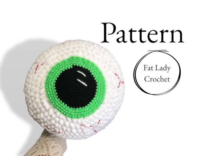 A giant crocheted eyeball with a green iris being held up against a white background with text that says "Pattern" and "Fat Lady Crochet."