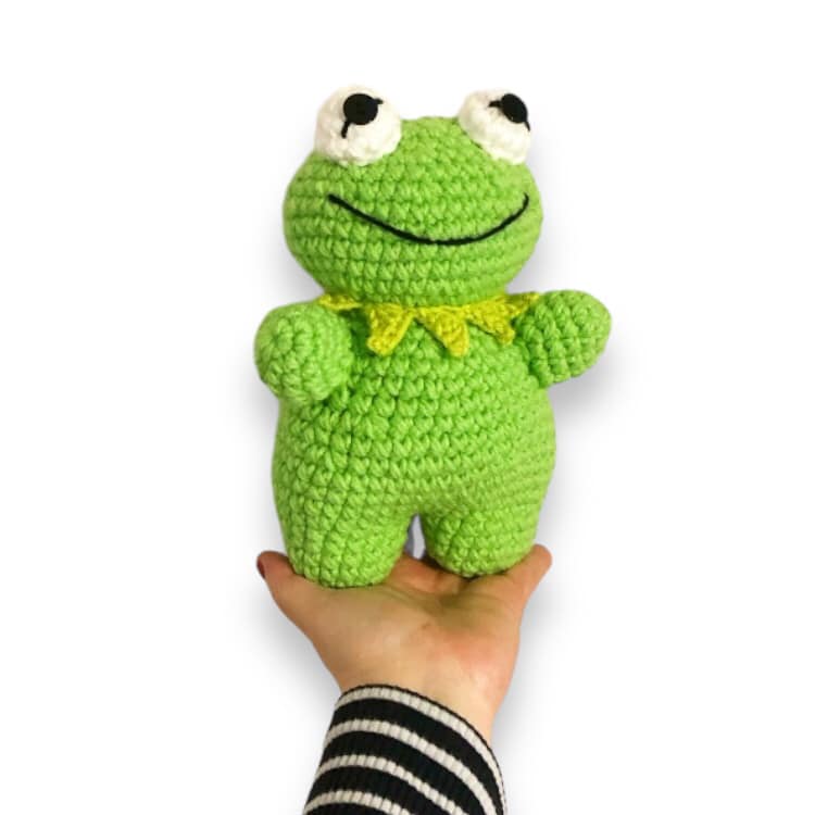 A crochet doll resembling Kermit being held up against a white background.