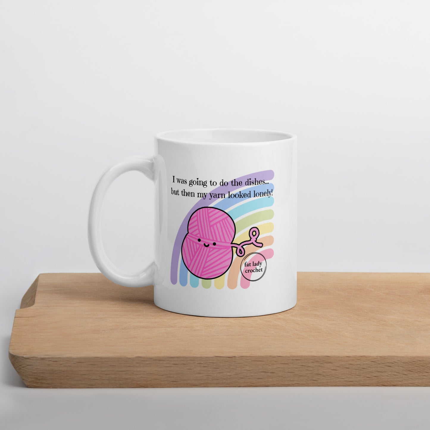 White mug with pink smiling yarn with a pastel rainbow set behind it set on a wooden board.