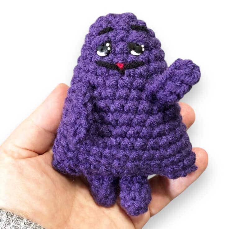A crocheted doll of Grimace being held up against a white background.