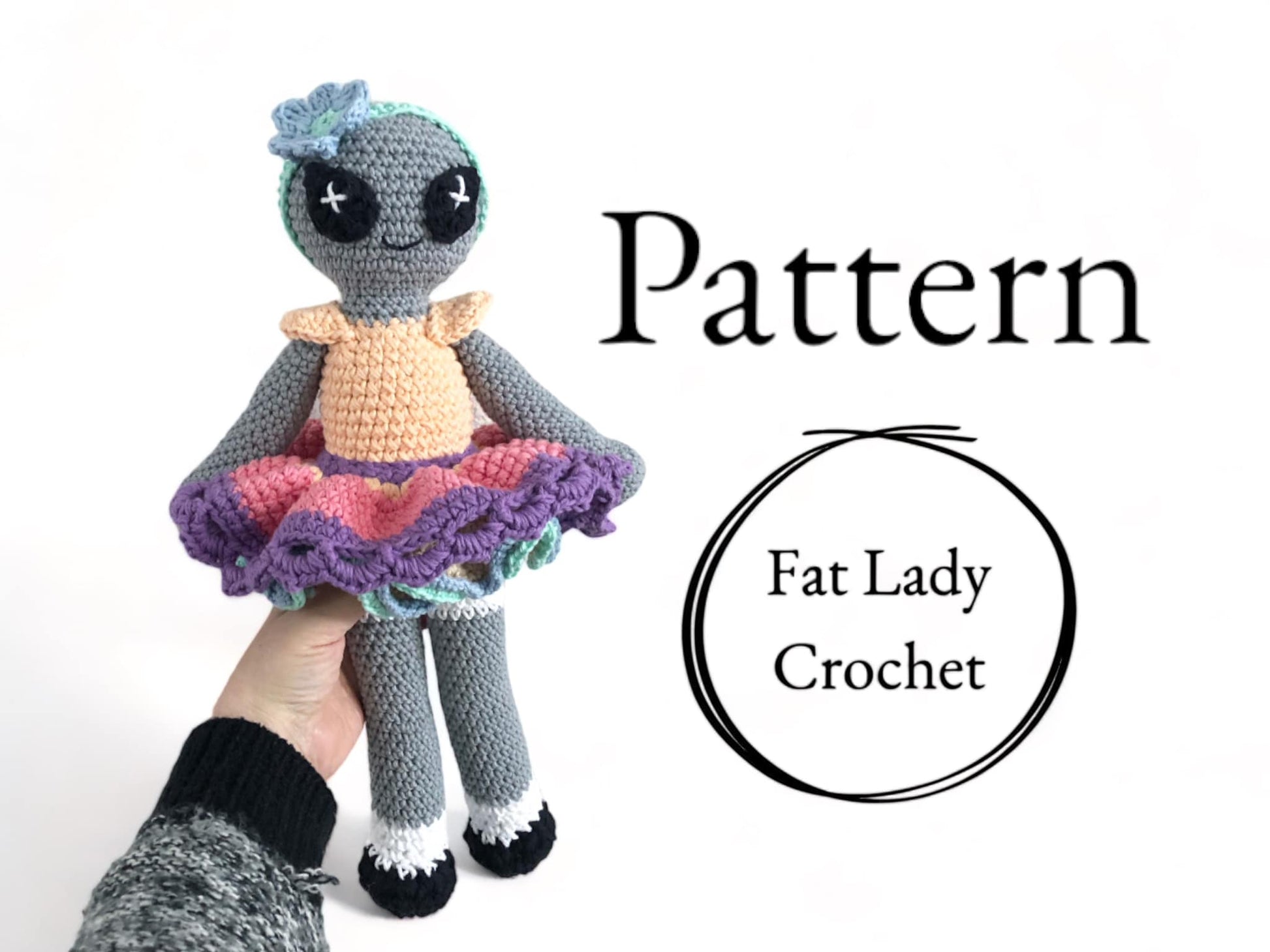 A crocheted doll of a colorful alien doll with a gray body, wearing a multicolored skirt and a pale orange top, held in a person's hand with text that reads "Pattern," and "Fat Lady Crochet."