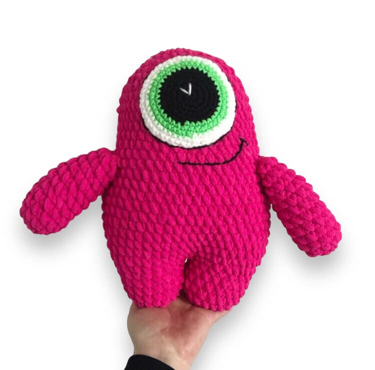 A crocheted pink monster with green eye plushie being held up against a white background