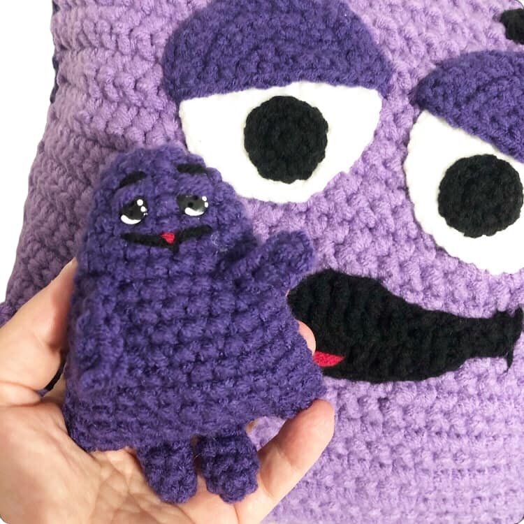 A crocheted doll of Grimace with a crochet Grimace pillow being in the background.