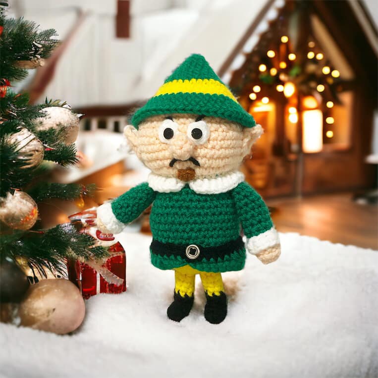 A crocheted version of the Head Elf from Rudolph the Red-Nosed Reindeer standing with some christmas decor.
