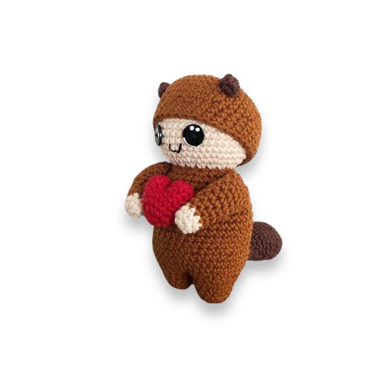 A crochet doll dressed in a brown squirrel costume holding a red heart.