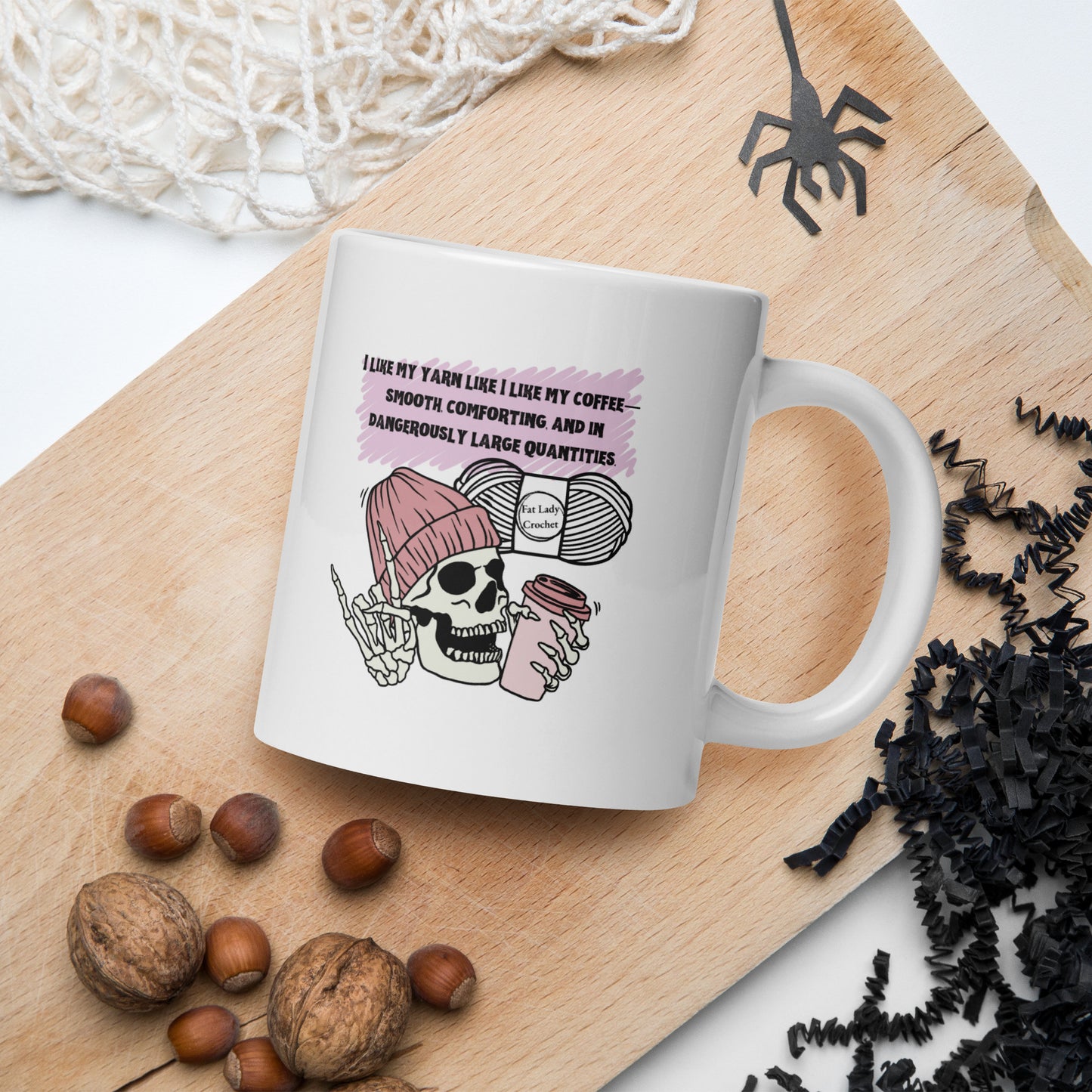 White mug with skeleton wearing a dark pink beanie holding a coffee mug with Halloween and autumn decorations surrounding it.