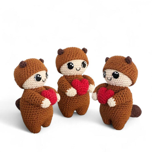 Three crochet dolls dressed like squirrels with brown bodies and beige faces, one holding a red heart, with black eyes and buttons, against a white background.