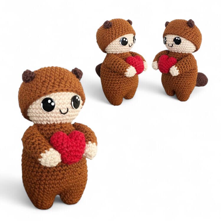Three crochet dolls dressed like squirrels with brown bodies and beige faces, one holding a red heart, with black eyes and buttons, against a white background, one of them brought forward into the foreground and the other two in the bakground.