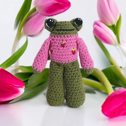 A crochet doll of a frog with a pink sweater with strawberries, displayed alongside pink tulips.