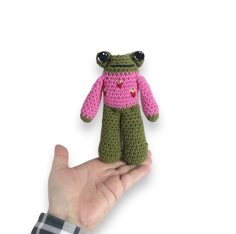 A crocheted frog doll in a pink sweater with strawberries being held up against a white background.