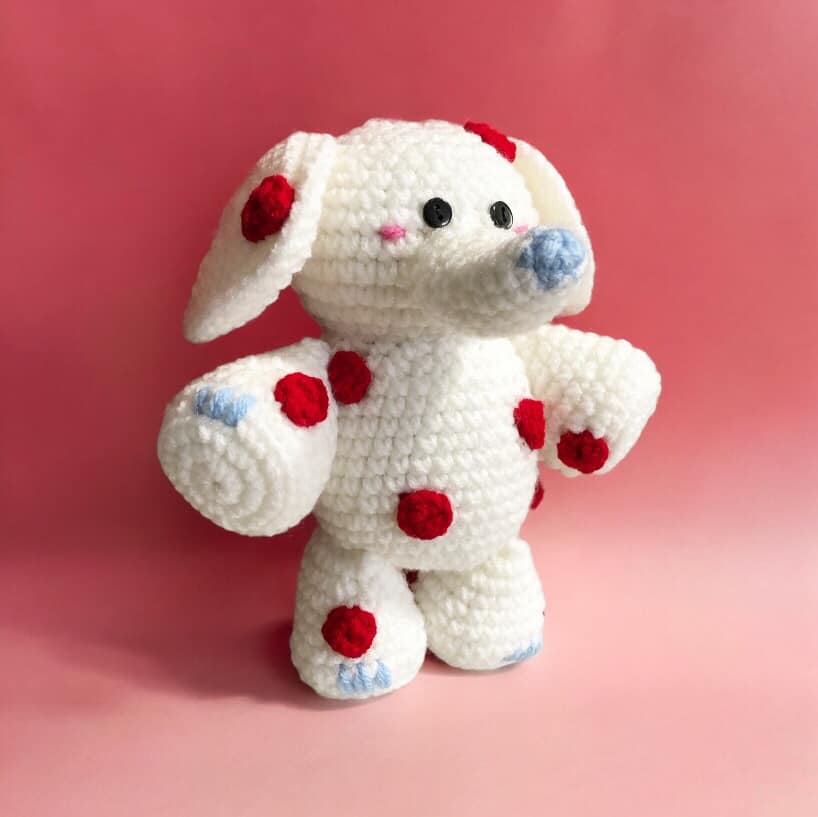 A crochet doll resembling the Spotted Elephant from Rudolph the Red-Nosed Reindeer standing against a pink background.