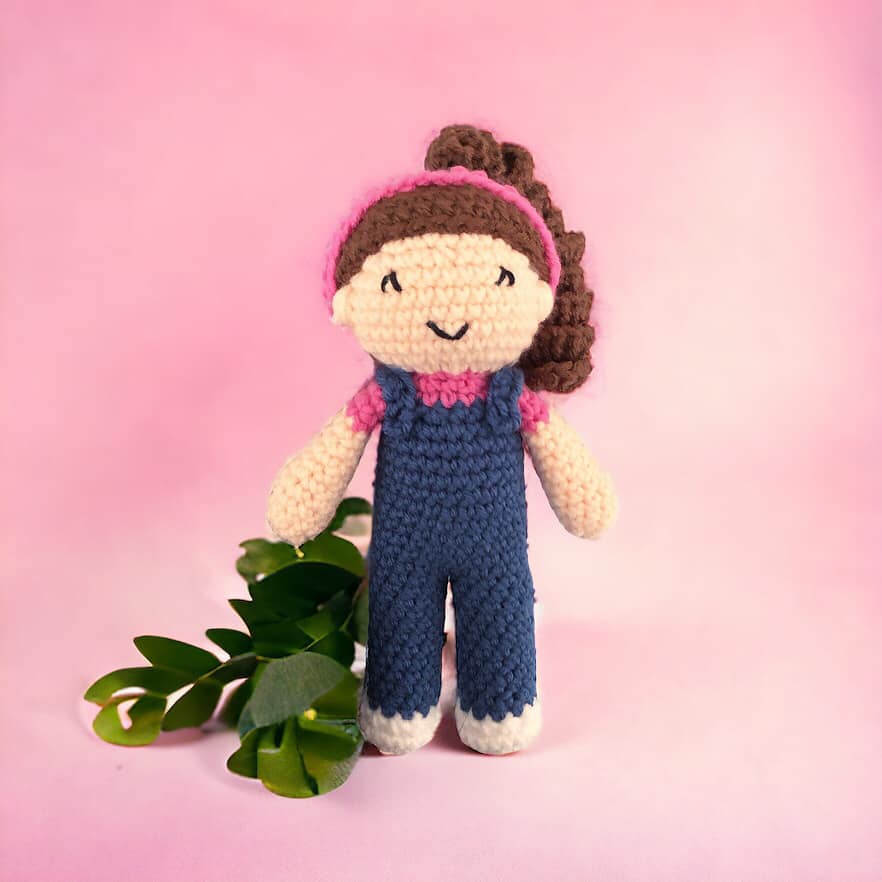 A crochet doll of a girl in overalls with a pink headband and a ponytail standing next to some leaves against a pink background