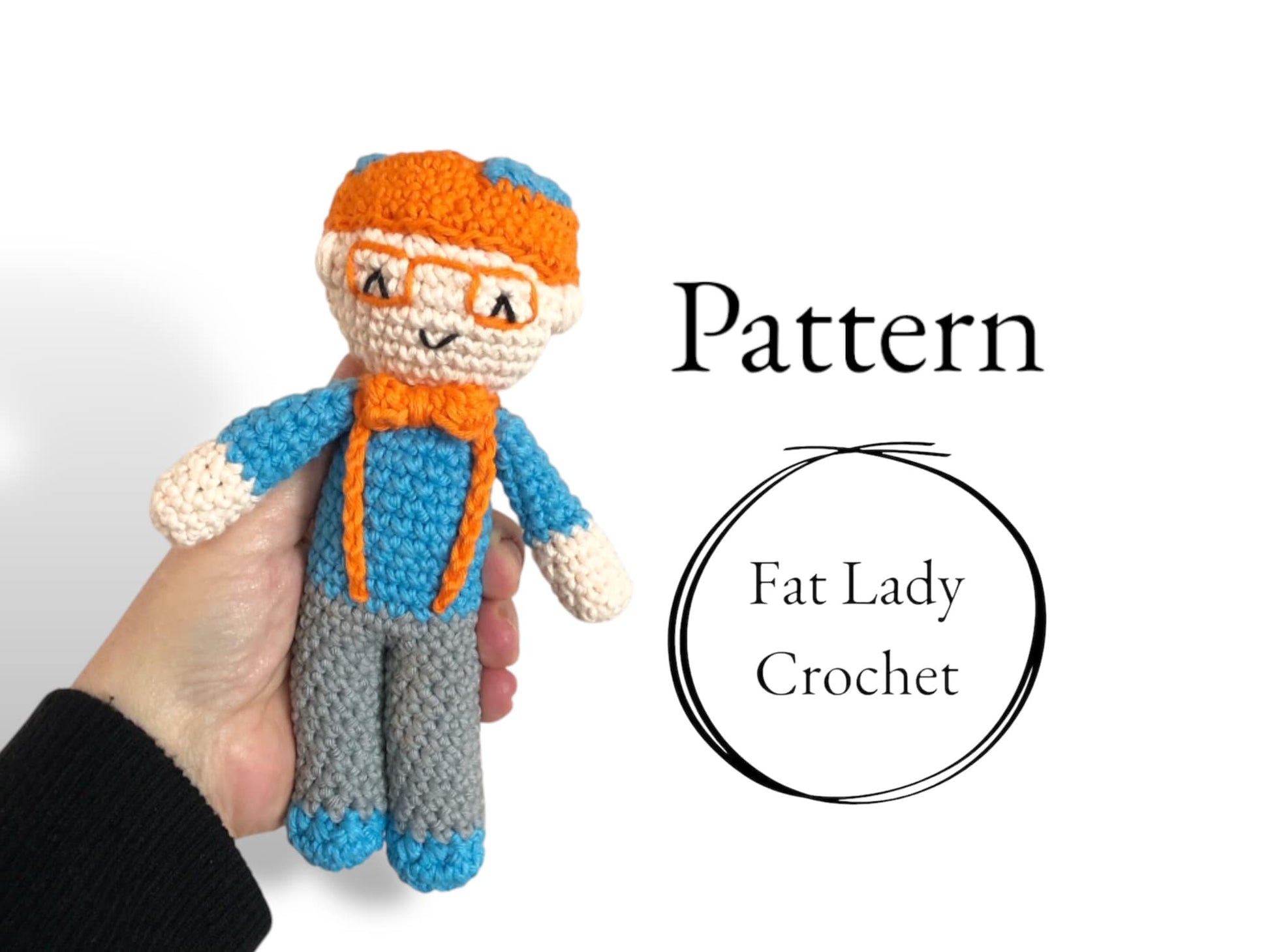One of the three crocheted dolls of the tiny tot trio, orange hair with orange glasses and a blue shirt, being held up against a white backdrop with the text "Pattern."