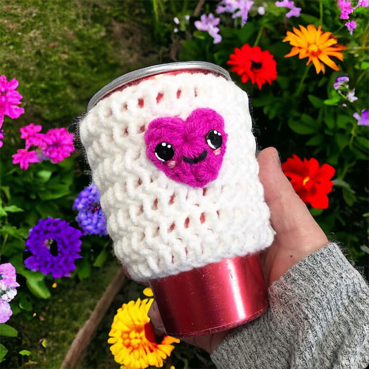 A crochet cup cozy with a heart pattern being held in front of a cup, displayed against a floral background.