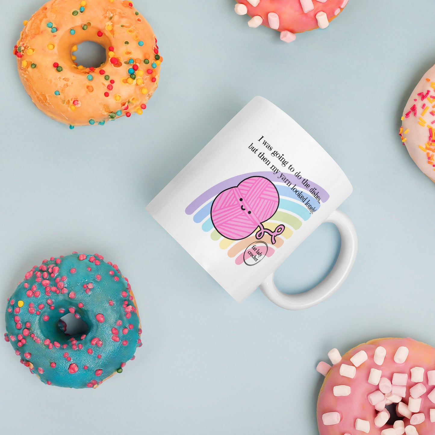 White mug with pink smiling yarn surrounded by very colourful donuts covered in sprinkles.