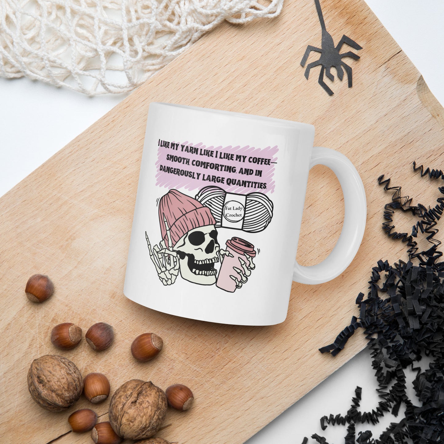 White mug with skeleton wearing a dark pink beanie holding a coffee mug with Halloween and autumn decorations surrounding it.