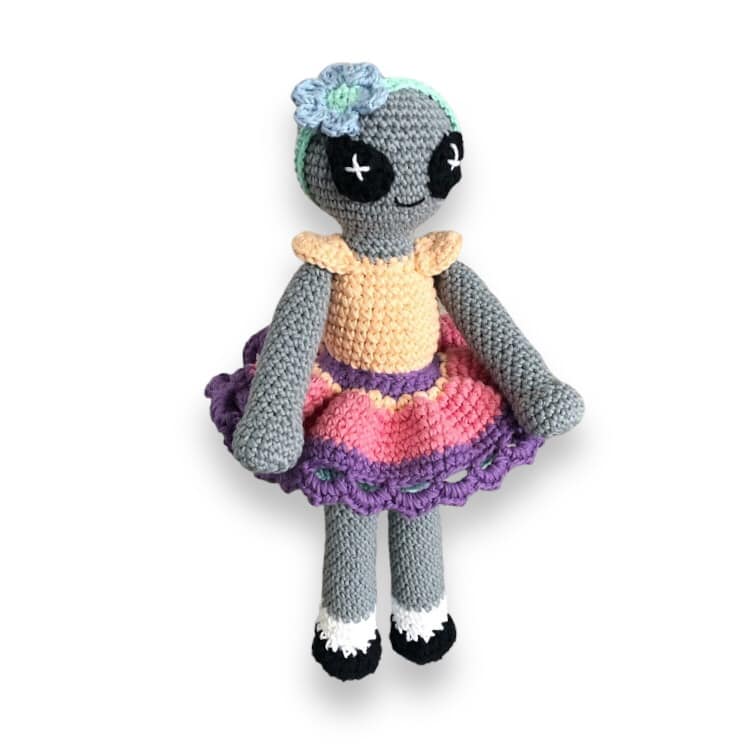 A crocheted doll of a colorful alien doll with a gray body, wearing a multicolored skirt and a pale orange top standing against a white background.