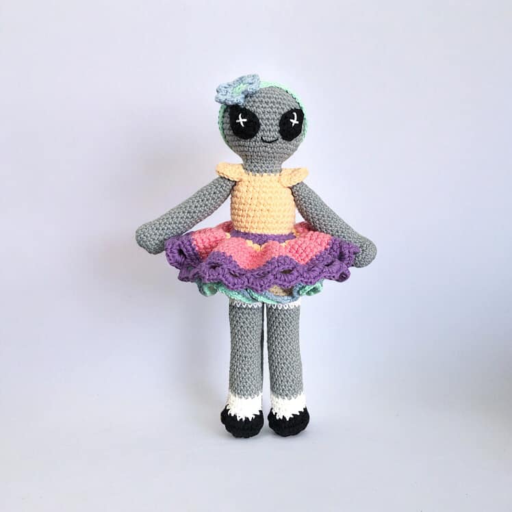 A crochet doll of an alien with a gray body, multicolored skirt, and decorative details.