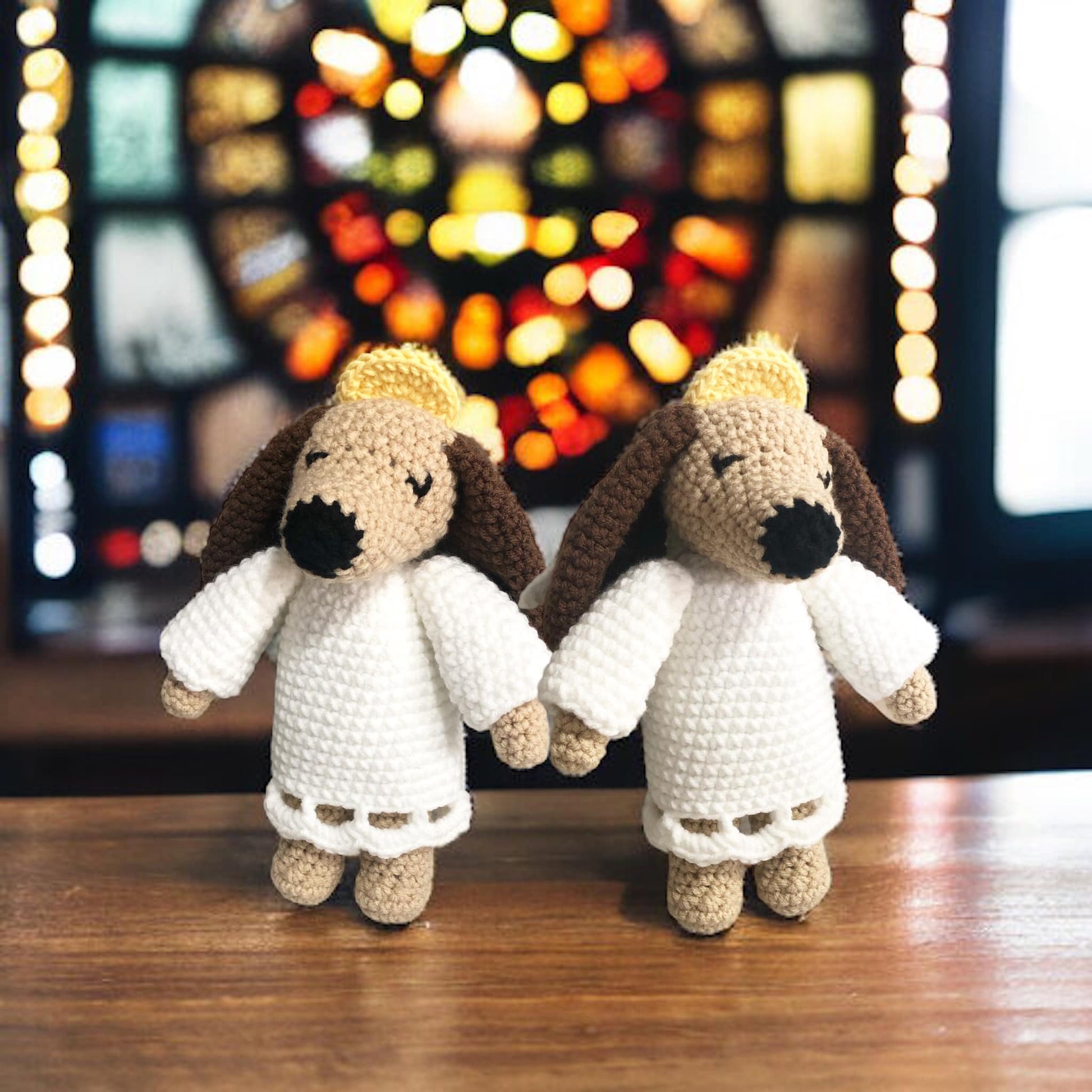 Two crocheted angel dog toys with beige, white, and brown colors, standing in front of a stained glass window.
