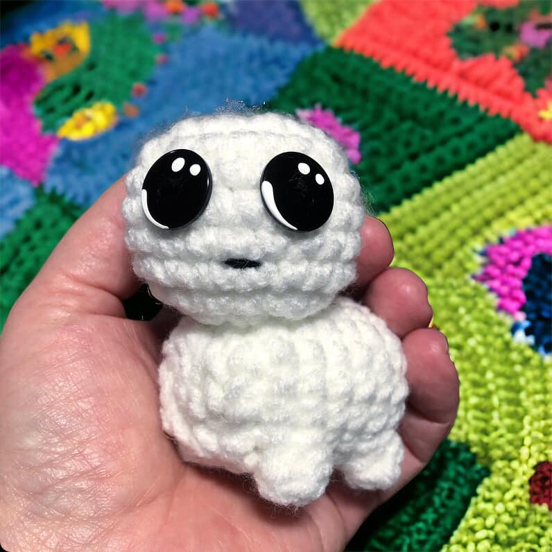 A crocheted version of the tbh creature being held up against a background of a knit blanket.