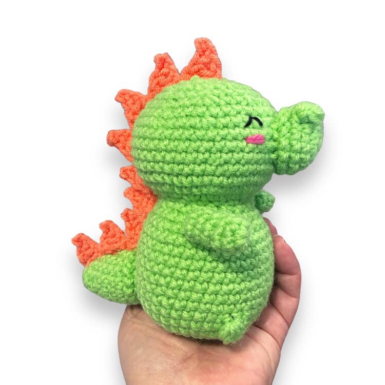 A small crochet baby dragon with a green body and orange spines, held in a person's hand.