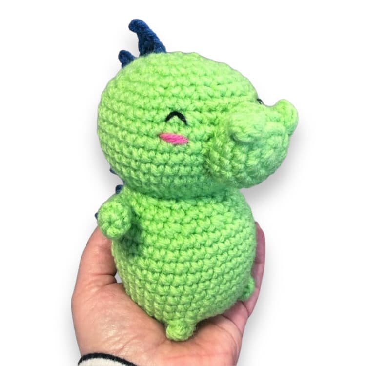 A small crochet baby dragon with a green body and blue spines, held in a person's hand.