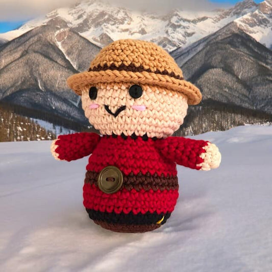A crochet doll resembling a Canadian Mountie standing on snow against a background of snowy mountains.